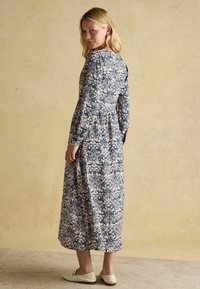 Sinine lilleline kleit pika varrukaga ja kogutud vöökoht, millel on kerge botaaniline muster, koos valgete libisemisega jalanõudega.