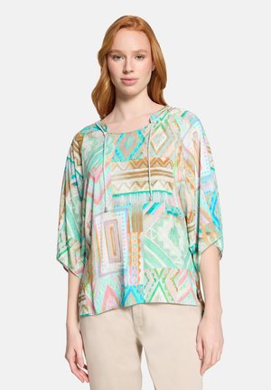 Rothaarige Frau, die eine locker sitzende Bluse mit pastellfarbenem geometrischem Muster und Zugbändern sowie beige Hosen trägt, steht vor einem schlichten Hintergrund.