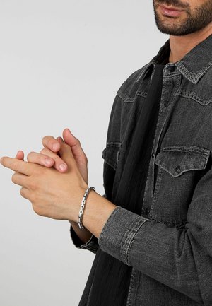 Camicia di denim grigia con bottoni a pressione, indossata sopra un tessuto nero. Al polso un bracciale a catena d'argento, composto da segmenti collegati.