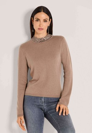 Frau in einem beigen Langarmpullover mit einem bestickten Kragen und blauen Jeans, die vor einem schlichten hellgrauen Hintergrund steht.