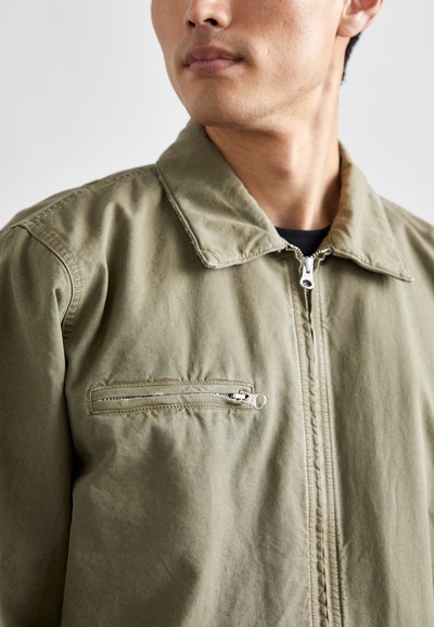 Olive green cotton jacket featuring a point collar, front zip closure, and a zippered breast pocket. Textured fabric with a relaxed fit.
