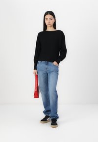 Zwarte gebreide trui met een ruime halslijn, gecombineerd met lichtblauwe rechte jeans en zwarte sneakers. Houdt een rode handtas vast.