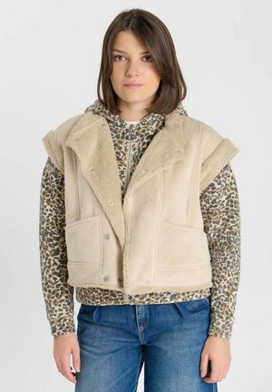 Junge Frau trägt eine beige ärmellose Fleecejacke über einem Leo-Print Reißverschluss-Top und blauen Jeans vor einem schlichten Hintergrund.
