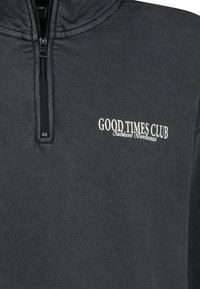 Zwarte zip-up sweatshirt met een hoge kraag. Heeft witte geborduurde tekst die "GOOD TIMES CLUB" zegt op de linkerborst. Zachte textuur.