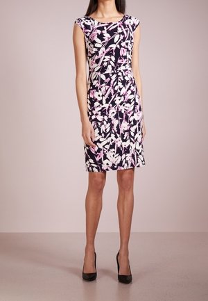 Woman wearing a knee-length sleeveless dress with a black, white, and pink floral pattern and black high-heeled shoes.