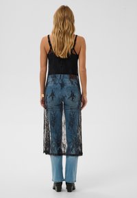 Schwarzes Oberteil mit Spitzenüberzug und dünnen Trägern, kombiniert mit blauen Jeans, die eine strukturierte Oberfläche, Gesäßtaschen und ein ausgestelltes Bein-Design aufweisen.