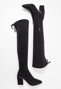 Schwarze Überknie-Stiefel aus glattem Stoff, mit spitzer Zehe, blockhaftem Absatz und dekorativen metallischen Nieten entlang der Sohle. Reißverschlussdetail an der Seite.