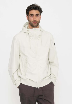 Uomo con capelli scuri e barba che indossa una giacca beige chiaro con cappuccio e cerniera e pantaloni scuri, in piedi con le mani nelle tasche su sfondo bianco.