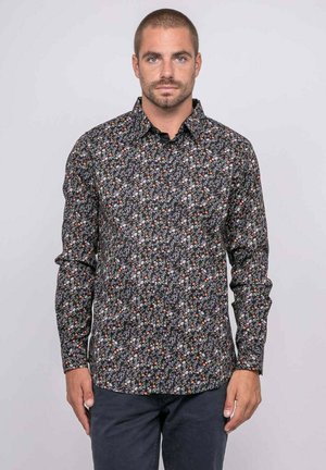 Homme portant une chemise noire à manches longues avec un petit motif floral en orange, blanc et violet, debout devant un fond uni gris clair.