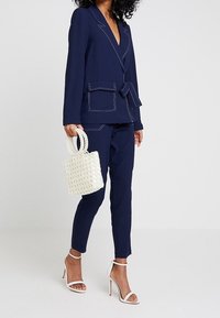 Žena v námořnickém modrém obleku s bílým prošitím, držící bílé korálkové kabelky a oblečená v bílých sandálech na vysokém podpatku.