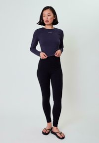 Donkerblauwe lange mouw top met een ronde hals en wit logo. Zwarte hoge taille leggings en zwarte slippers maken de outfit compleet.