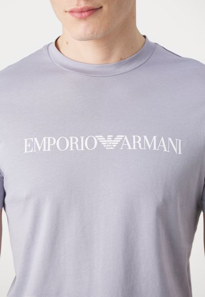 Heled lilla puuvillane T-särk, millel on valge "EMPORIO ARMANI" logo. Kujundus sisaldab logol iseloomulikku kotka embleemi.