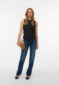 Sieviete stāv, ģērbta melnajā piedurkņu krekliņā, zilās džinsās un melnos smailos apavos, tur rokā brūnu rokassomiņu uz vienkārša baltā fona.