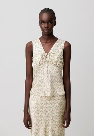 Donna con i capelli intrecciati che indossa un top crema senza maniche e una gonna abbinata con motivi floreali geometrici verdi e blu.