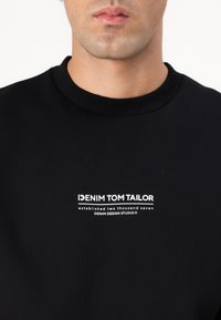 Schwarzer Kapuzenpullover mit rundem Ausschnitt, der mit weißem Drucktext "DENIM TOM TAILOR" versehen ist, aus weichem, strukturiertem Stoff gefertigt.