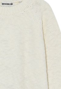 Ljusgrå stickad tröja med ribbad rundhals, raglanärmar och ett texturerat mönster. Lappen säger "MINIMUM."