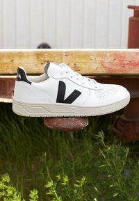 Valkoinen nahkakengät mustilla ja harmailla yksityiskohdilla, rei'itetty kynnen osa, pyöreä kärkimuoto ja tekstuuri vaaleanbeige pohja. Brändilogo kantapäässä.