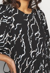 Svart klänning med ett vågigt vitt och beige mönster, med puffärm och ett mjukt, texturerat tyg. Lätt insydd under bysten.