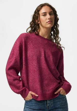 Femme aux cheveux bouclés portant un pull violet ample à manches longues et un jean bleu, debout les mains dans les poches devant un fond uni.