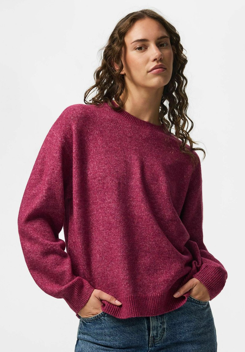 Femme aux cheveux bouclés portant un pull violet ample à manches longues et un jean bleu, debout les mains dans les poches devant un fond uni.