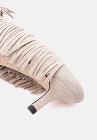 Chaussure beige en suède à talon kitten avec des détails à franges superposées et une petite pointe de talon noire, vue de profil sur fond blanc.