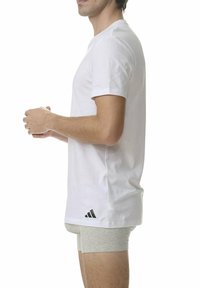 Mężczyzna w prostym białym t-shircie Adidas i szarych szortach, stojący w profilu z rękami splatanymi, na białym tle.