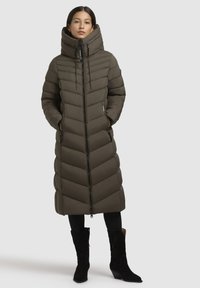 Polstret lang frakke i olivengrøn med et chevronmønster, høj krave, lynlås foran og sidelommer; parret med sorte ankelstøvler.