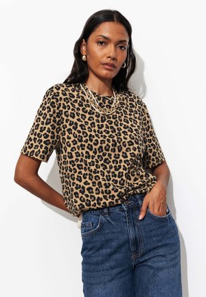 Femme aux longs cheveux foncés portant une chemise à manches courtes imprimé léopard, un jean bleu, un collier chaîne en or et des boucles d'oreilles en or, debout sur fond blanc.