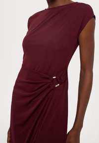 Burgunderfärgad klänning med draperad design, utrustad med en knuten detalj som säkras med ett guldcirkelfäste. Slät, elastisk tyg.