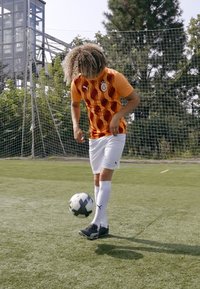 Maglietta da calcio a righe arancioni e rosse con logo, pantaloni bianchi, scarpini neri; giocatore che dribbla un pallone da calcio bianco e nero su un campo verde.