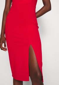 Kobieta w dopasowanej czerwonej sukience z wysokim rozcięciem na lewym boku na jednolitym jasnym tle.