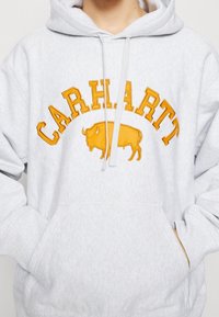 Hellegrauer Hoodie mit gelbem "CARHARTT"-Text und Büffel-Logo, die Hände in der vorderen Tasche verstaut.