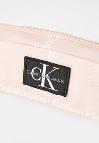 Rosa tygpenna med en svart etikett som visar logotypen "CK" och "Calvin Klein Jeans", med dragkedjestängning högst upp, texturerat material.