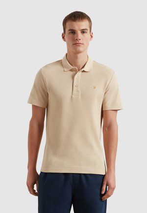 Beige geripptes Poloshirt mit einer zweiknöpfigen Knopfleiste und kurzen Ärmeln. Mit einem auf der Brust gestickten Logo. Kombiniert mit dunkelblauen Shorts.