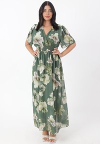 Robe maxi florale en tissu vert avec des manches bouffantes, une taille cintrée et une jupe fluide ornée de grandes fleurs blanches et violettes.
