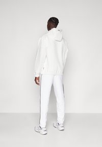 Vit huva jacka med texturerad mönster, elastiska ärmar och dragsko. Sammanförd med vita byxor som har en svart sidostrip och vita sneakers.