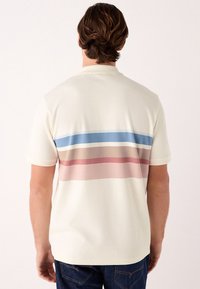 T-shirt color crema con un disegno sulla schiena caratterizzato da strisce orizzontali in blu, beige, rosa e rosso scuro. Maniche corte e tessuto morbido.