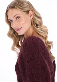 Bordeaux gebreide trui met een losse pasvorm en textuurpatronen, gecombineerd met gouden oorbellen met strass-accenten en golvend blond haar.