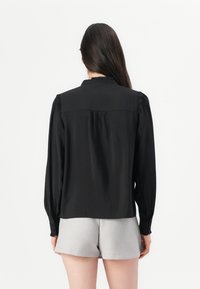 Blusa nera con maniche lunghe, scollatura arricciata e pieghe posteriori. Tessuto liscio, abbinata a pantaloni corti grigi chiari per un look casual.