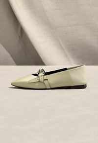 Chaussure plate crème à bout pointu avec une double bride à boucle argentée et une semelle basse noire, sur un fond en tissu beige clair.