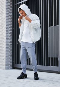 Λευκό μπουφάν με κουκούλα, λεπτομέρειες από δίχτυ, γκρι φόρμα με λογότυπο και μαύρα αθλητικά, τοποθετημένα μπροστά σε μοντέρνο τοίχο από τούβλα και πύλη.