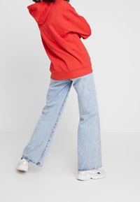 Person i ljusblå vida jeans, vita sneakers och en röd huvtröja står med armarna i kors, vänd något bort från kameran mot en neutral bakgrund.