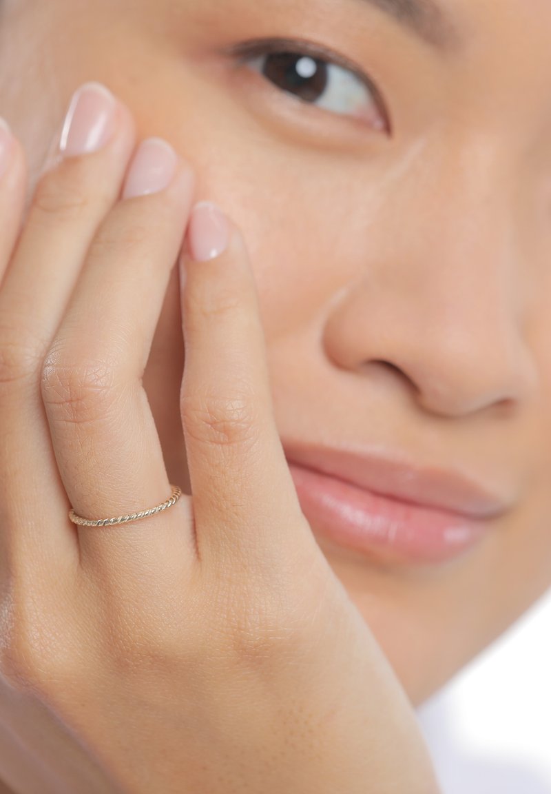 Elegante anello d'oro con un design intrecciato, indossato su una mano che tocca delicatamente il viso. Pelle liscia e colore neutro delle unghie sono visibili.