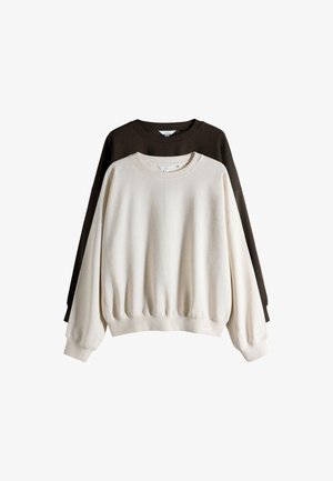 Jeune femme aux cheveux bruns portant un sweat-shirt beige oversize et un pantalon, assise en tailleur sur un sol clair devant un fond gris uni.
