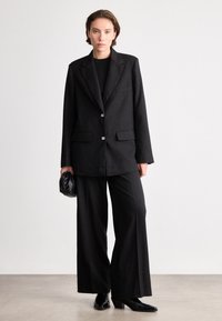 Sacou negru, ajustat, cu două butoane, rever înclinat și buzunare laterale, purtat cu pantaloni evazați. Țesătură fină, cu detalii texturale minime.