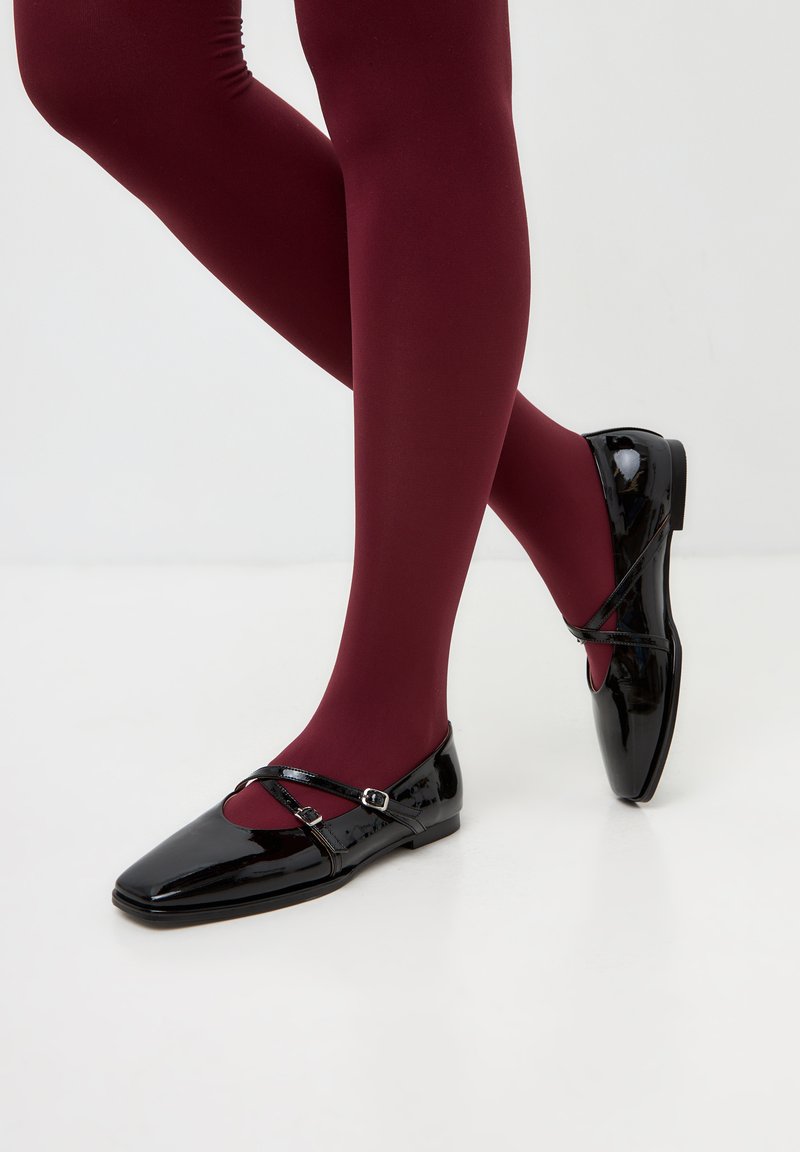 Zwarte gepatineerde ballerina's met een vierkante neus, voorzien van een kruislings strap en een gespsluiting, aangevuld met bordeauxrode panty’s.