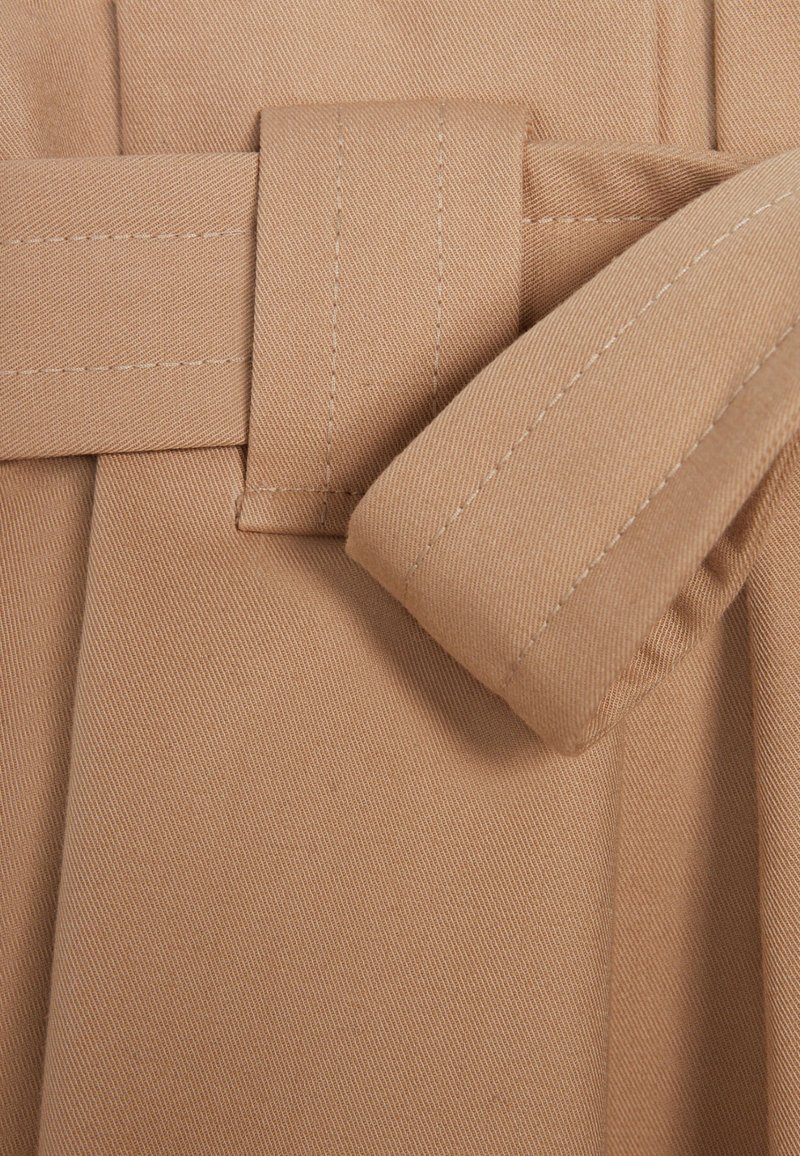 Detailaufnahme einer beigen Stoffgürtelsschlaufe mit einem durchgezogenen genähten Gürtel, der die detaillierte Naht und Textur des Materials zeigt.