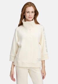 Sweat-shirt couleur crème avec col montant, poignets côtelés et fermeture par boutons. Détail texturé le long des manches ajoute un intérêt visuel.