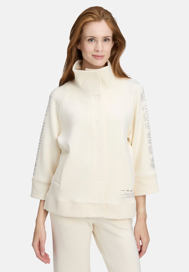 Sweat-shirt couleur crème avec col montant, poignets côtelés et fermeture par boutons. Détail texturé le long des manches ajoute un intérêt visuel.