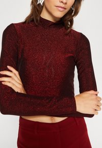 Rotes, glitzerndes Langarm-Crop-Top mit hohem Kragen und figurbetontem Schnitt, kombiniert mit passenden hochtaillierten Hosen.